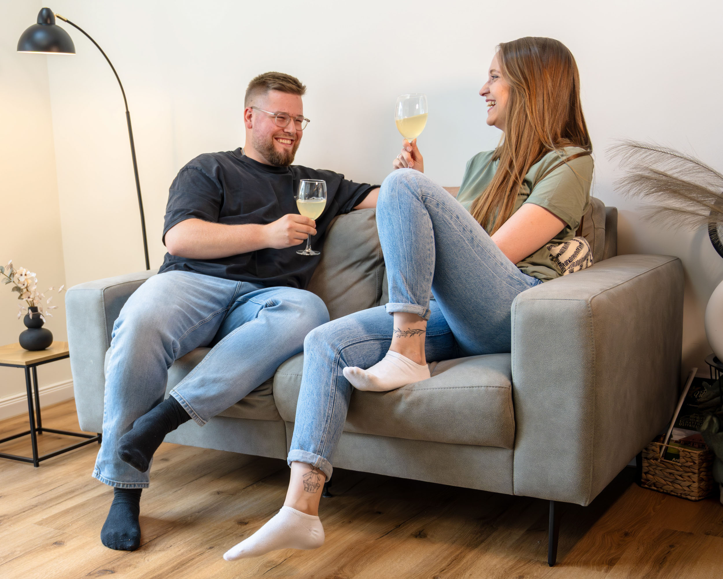 Junge Frau in Latzhose kniet auf dem Boden und hält lächelnd ein helles Laminatbrett in einem renovierungsbedürftigen Raum